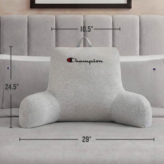 A Champion Jersey Backrest Pillow in gray from Champion rests comfortably on a bed, its arms providing support. The dimensions—24.5 inches tall, 29 inches wide, and 10.5 inches deep—are clearly noted. This cozy pillow is adorned with the iconic Champion logo and placed against a tufted headboard.