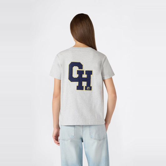 A woman with long brown hair is wearing a relaxed fit light gray Champion Women’s Knitted Crewneck T-Shirt, featuring large blue and yellow “CH” letters on the back, paired with light blue jeans, while standing against a plain white background.