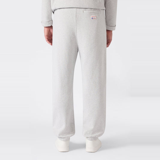 A person is shown from the back wearing Men’s Knitted Elastic Cuff Pants by Champion in light gray, paired with a matching sweatshirt. These trousers feature a loose fit, elastic cuffs, and a small logo patch on the right side near the waistband. Crafted from heavyweight fleece for comfort, the set stands out against a plain white background.