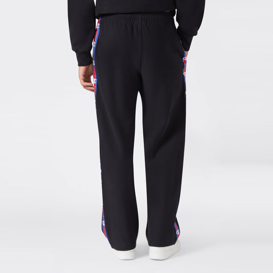 Back view of a person wearing black Champion Men’s Knitted Straight Hem Pants featuring red, white, and blue logo tape along the sides, paired with a matching black sweatshirt and white sneakers against a plain light gray background—an ideal option for men’s sportswear.