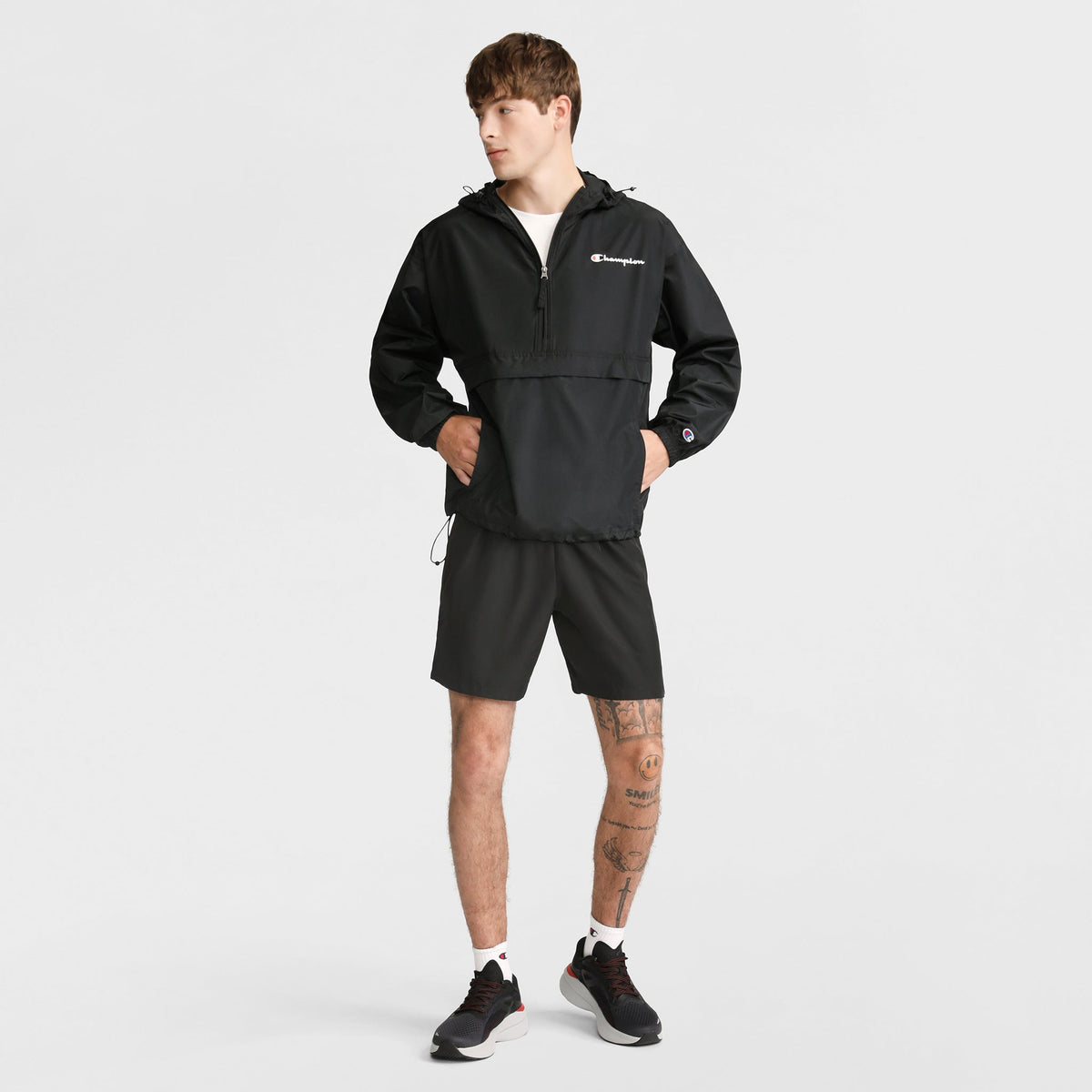 A person stands against a white background, wearing a black Champion jacket, Champion 7" Woven Sport Shorts, white socks, and black sneakers. They have tattoos on one leg and look to the side with hands in their jacket pockets.