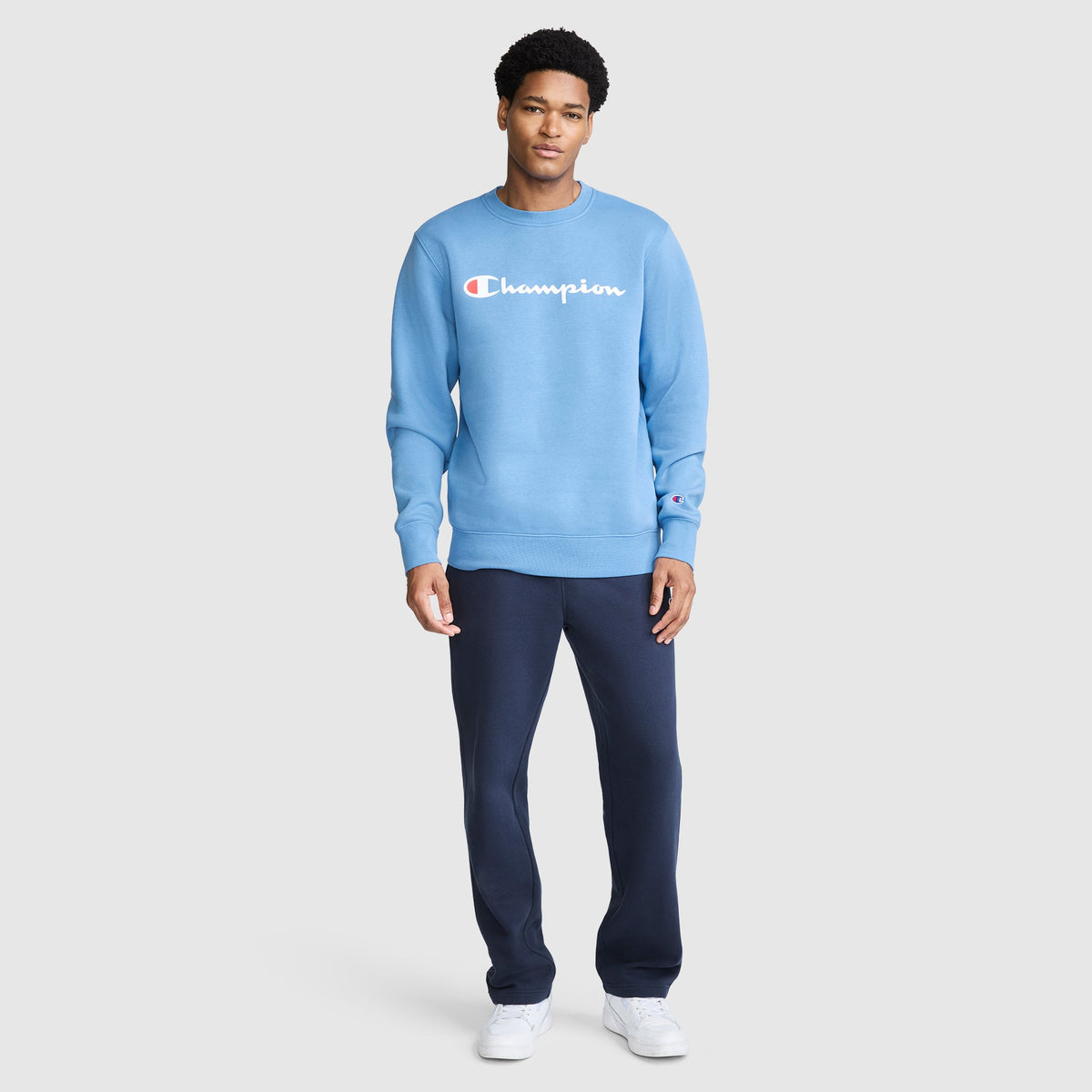 A man stands facing forward, wearing a light blue Champion Powerblend Crewneck Sweatshirt with Classic Script Logo, dark blue pants, and white sneakers, against a plain light gray background.