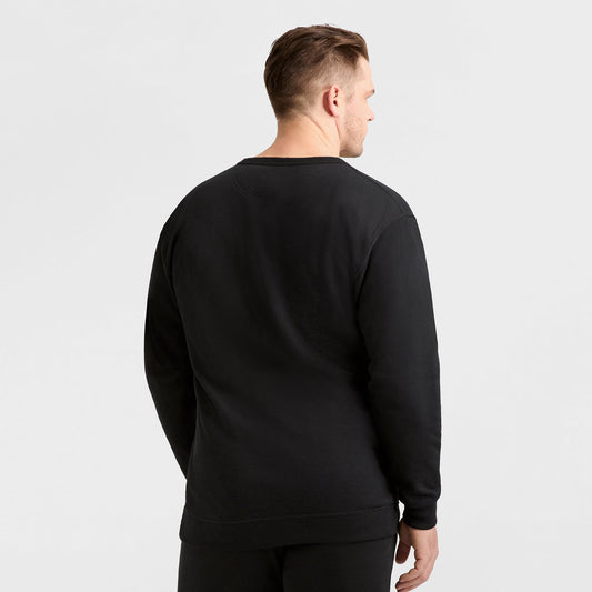 A man with short hair, facing away from the camera, is wearing a plain black Champion Powerblend Crewneck Sweatshirt, C Logo - Big & Tall, and black pants against a light gray background.