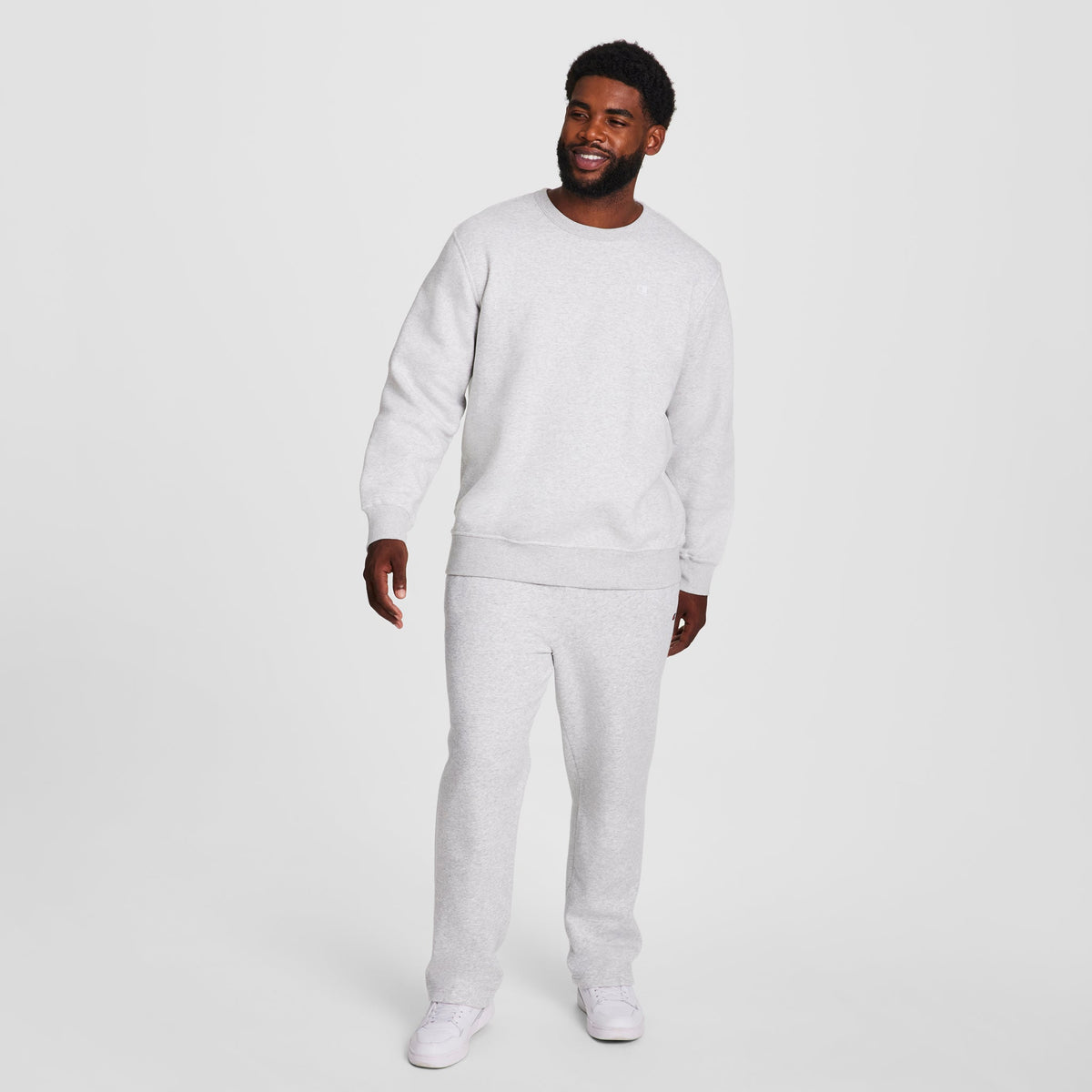 A man smiles wearing a Champion Powerblend Crewneck Sweatshirt, C Logo - Big & Tall, paired with matching sweatpants and white sneakers, standing against a plain white background.