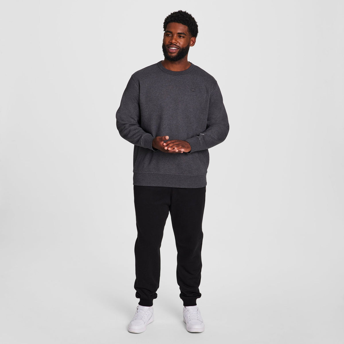 A man smiles against a white background, wearing the Champion Powerblend Crewneck Sweatshirt, C Logo - Big & Tall in dark gray, paired with black sweatpants and white sneakers. He clasps his hands, highlighting the cozy style.
