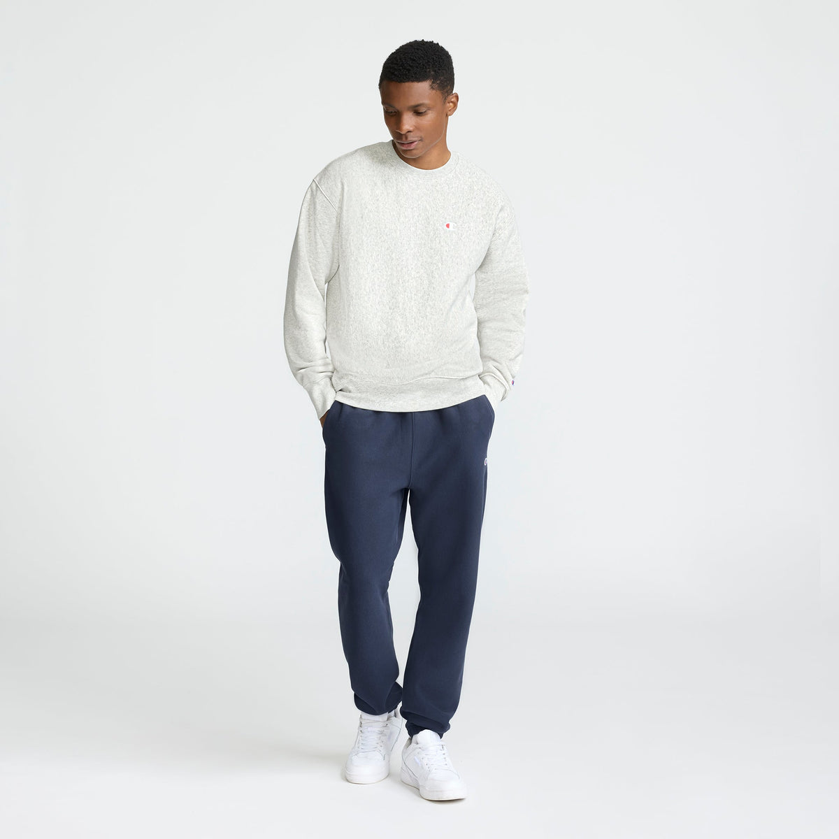 A man stands with his hands in his pockets, looking down in a minimalist white studio, wearing a Champion Reverse Weave Crewneck Sweatshirt with the C Logo, dark blue jogger pants, and white sneakers.