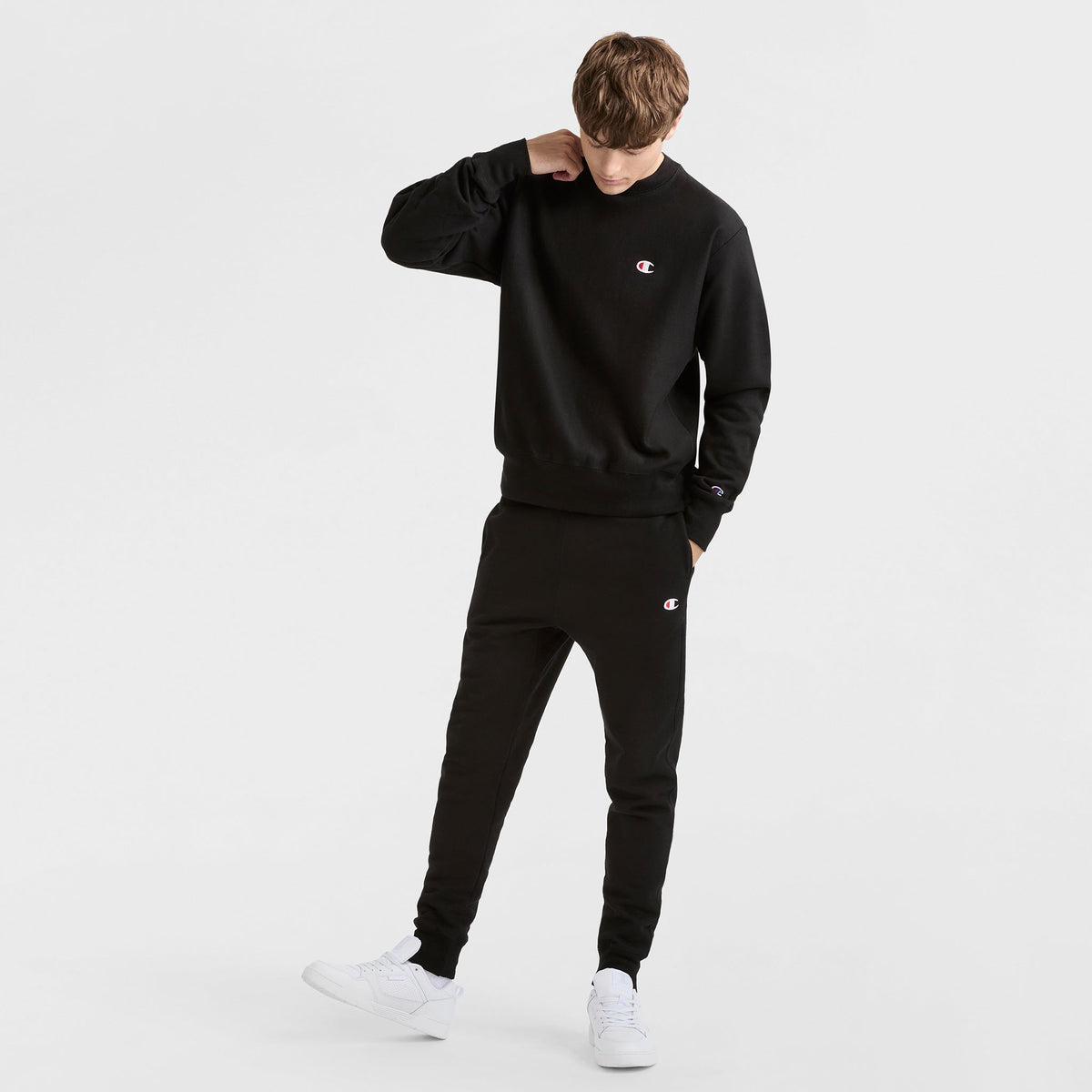 A person wearing a Champion Reverse Weave Crewneck Sweatshirt, C Logo, and matching black sweatpants with white sneakers stands against a plain white background, looking down with one hand touching their neck.