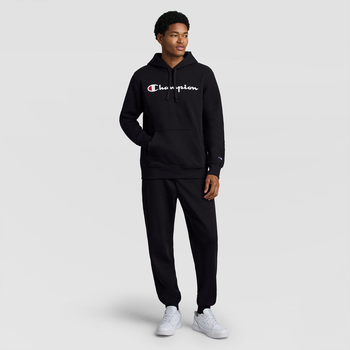 A person wearing the Champion Powerblend Hoodie, Full Script Logo in black with matching sweatpants stands smiling with one hand in the hoodie pocket, paired with white sneakers, against a plain light gray background.