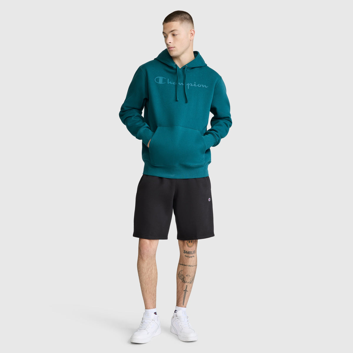 A person wearing a teal Champion Powerblend Hoodie with a tonal script logo pairs it with black shorts and white sneakers, standing against a plain light background and looking to the side, hands in the hoodie’s front pocket.
