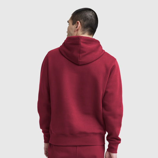 A person wearing a maroon Champion Powerblend Hoodie, C Logo and matching pants stands with their back to the camera against a plain, light background, looking slightly to the left.