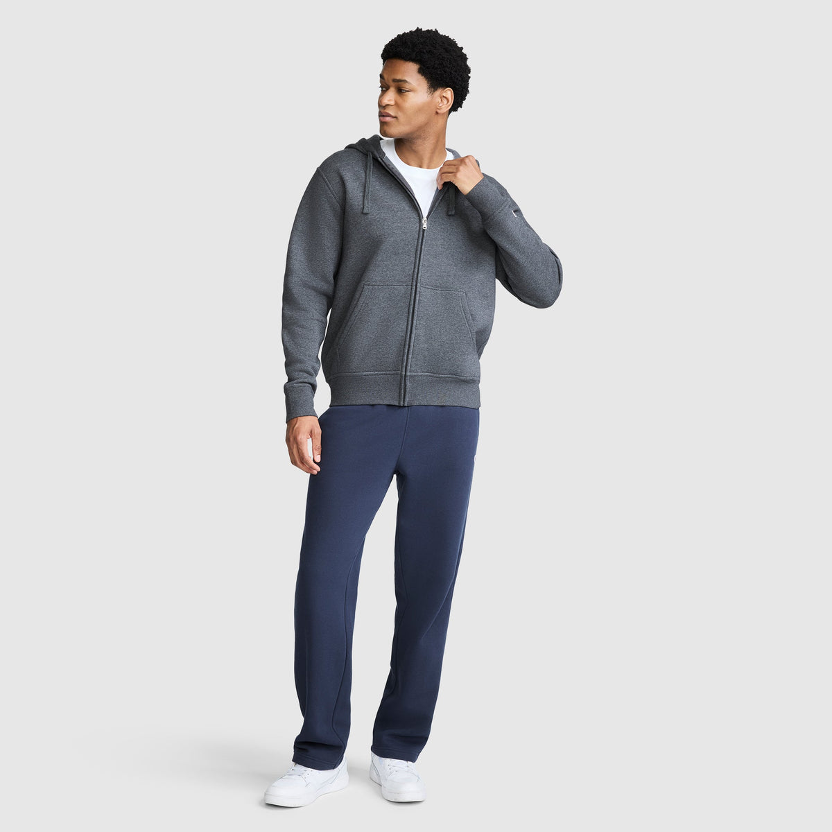 A man wearing a gray Champion Powerblend Full-Zip Hoodie, white t-shirt, navy blue Champion men's sweatpants with Powerblend cinched bottoms, and white sneakers stands against a plain light gray background, looking to his left with one hand on his collar.