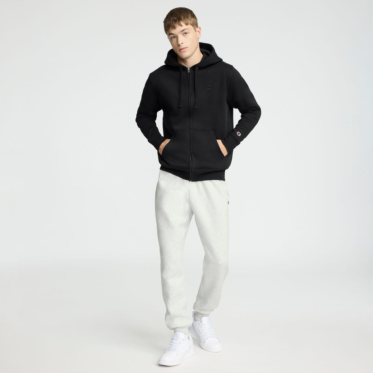 A person stands facing forward wearing a Champion Powerblend Full-Zip Hoodie, C Logo—a black men’s hoodie—with light gray sweatpants and white sneakers against a plain white background. Their hands are in the hoodie pockets.