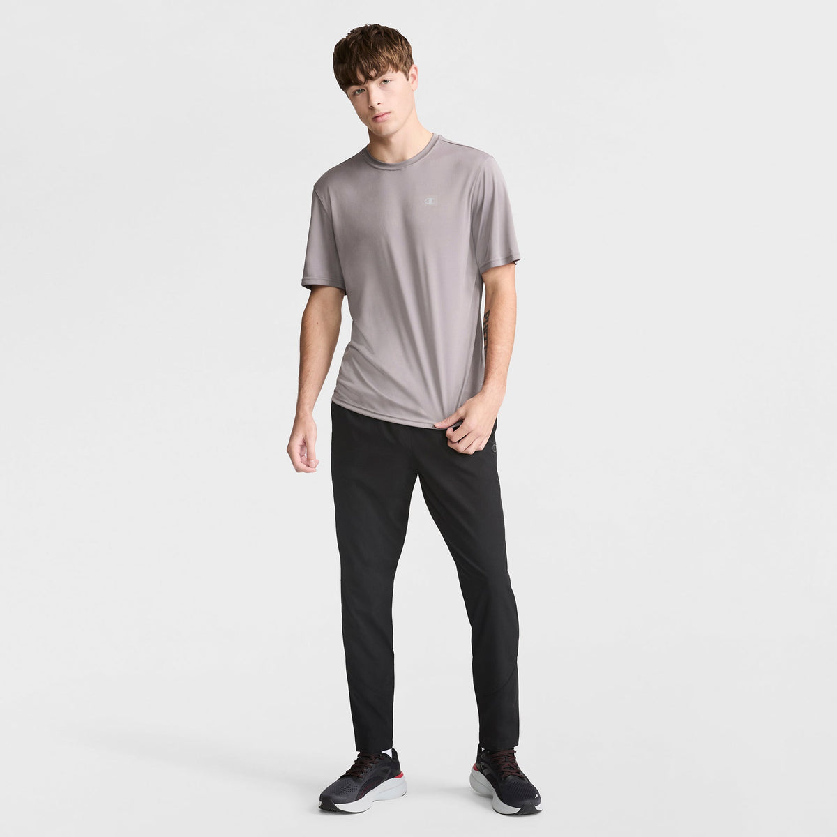 A young man stands against a white background, wearing Champion MVP Pants with moisture wicking and C Logo, paired with a light gray athletic shirt and black sneakers. He has short brown hair and a relaxed posture.