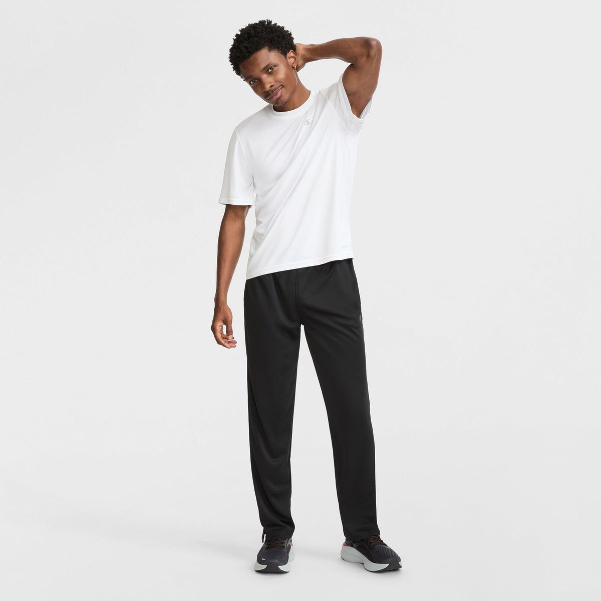 A man stands against a white background wearing black Champion Sport Sweatpants with the C logo, a lightweight white short-sleeve athletic shirt, and black sneakers. He smiles, looking slightly to the side with one hand behind his neck.