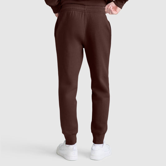 A person is shown from the back wearing Champion Powerblend Joggers, C Logo in brown with white sneakers, standing against a plain light gray background.