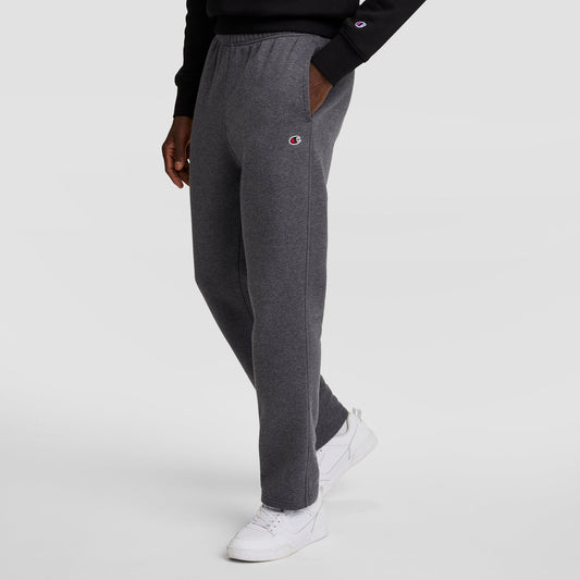 A person in dark gray Champion Powerblend Sweatpants (Open Hem, C Logo), a black sweatshirt, and white sneakers stands against a light gray background. The Champion logo is visible on the sweatpants and sweatshirt sleeve.