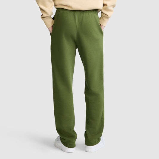 The person is wearing olive green Champion Powerblend Sweatpants, Relaxed Bottom, C Logo, made from a soft cotton blend. The sweatpants are paired with a beige sweatshirt and white sneakers. Standing with hands in pockets against a plain white background, only the lower half of the body is visible from the back.
