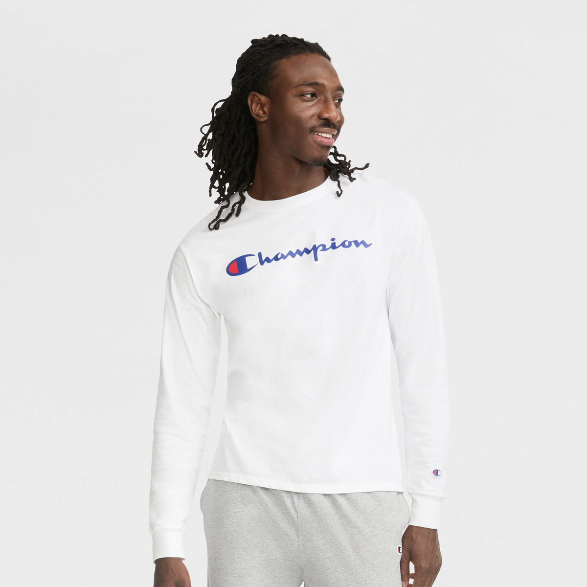 A man with long, braided hair wears a white Champion shirt and Champion Lightweight Lounge Joggers with C Logo in light gray, smiling and looking to the side against a plain white background.