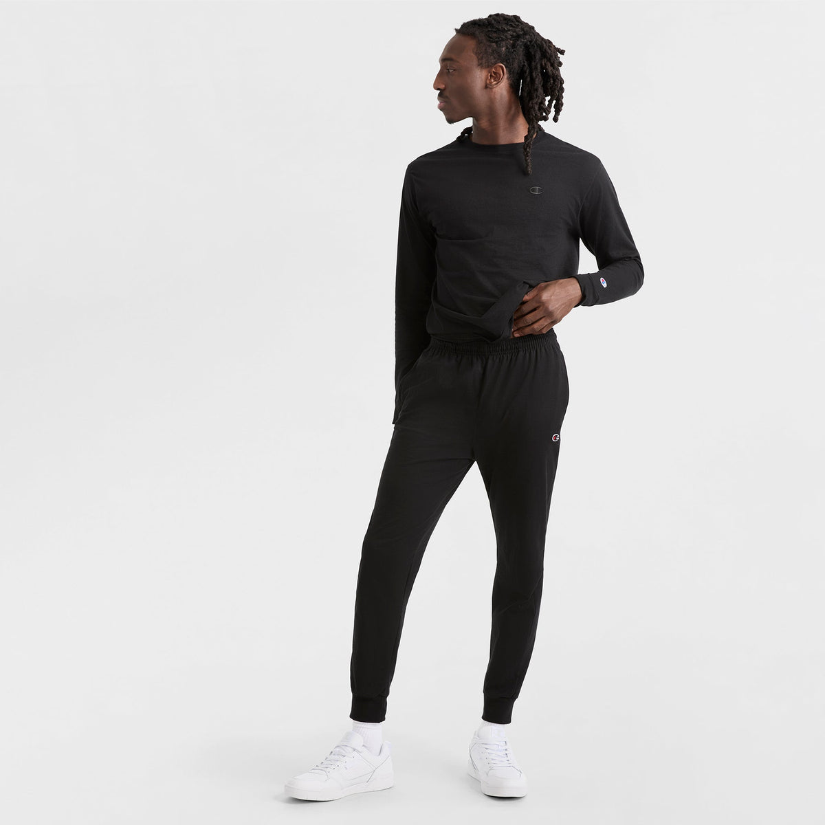 A person stands against a white background wearing Champion’s Lightweight Lounge Joggers, C Logo, featuring an elastic drawcord waistband, paired with a black long-sleeve shirt and white sneakers, looking to the side with one hand in their pocket.