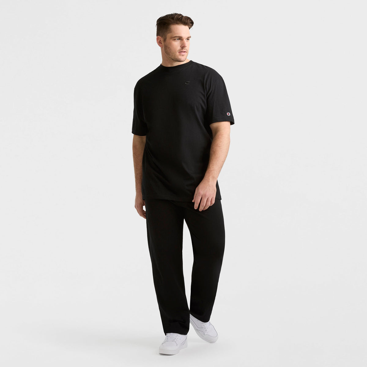 A man stands against a plain white background wearing a black t-shirt, Champion Lightweight Lounge Pants with an open hem and C logo (Big & Tall), and white sneakers. He looks slightly to the side, one hand by his side and the other near his hip.