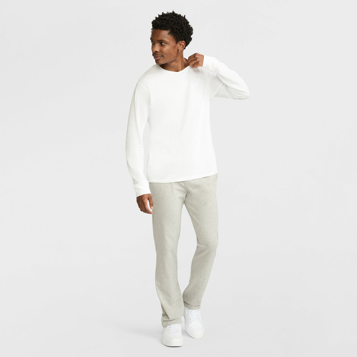 A man in Champion Lightweight Lounge Pants, Open Hem, C Logo, a white long-sleeve shirt, and white sneakers stands against a plain white background, looking left with one hand near his collar. The jersey knit adds casual comfort.