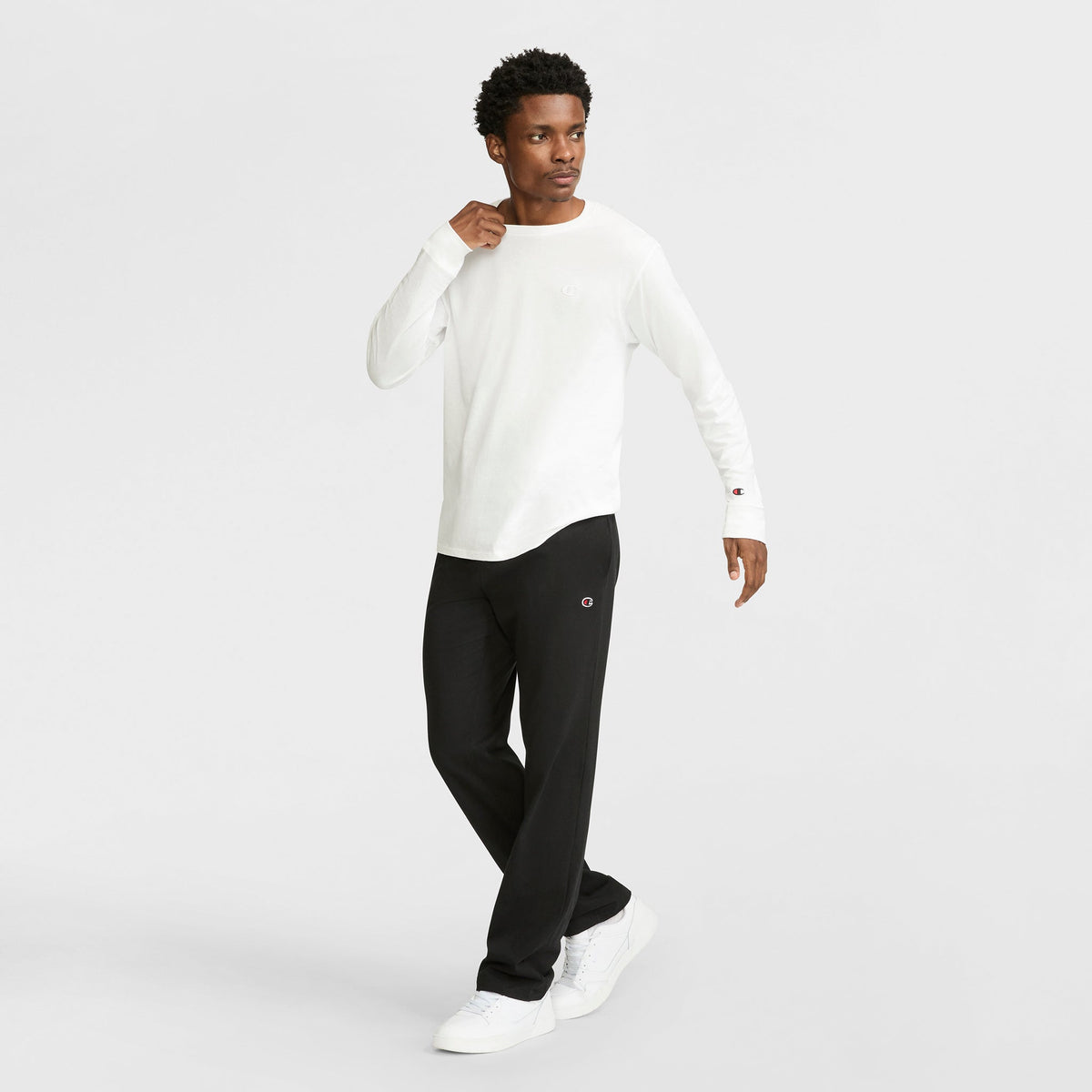 A man in a white Champion jersey knit long-sleeve shirt, black Champion Lightweight Lounge Pants with open hem and C Logo, and white sneakers stands against a plain light background, looking to the side with one hand near his chest.