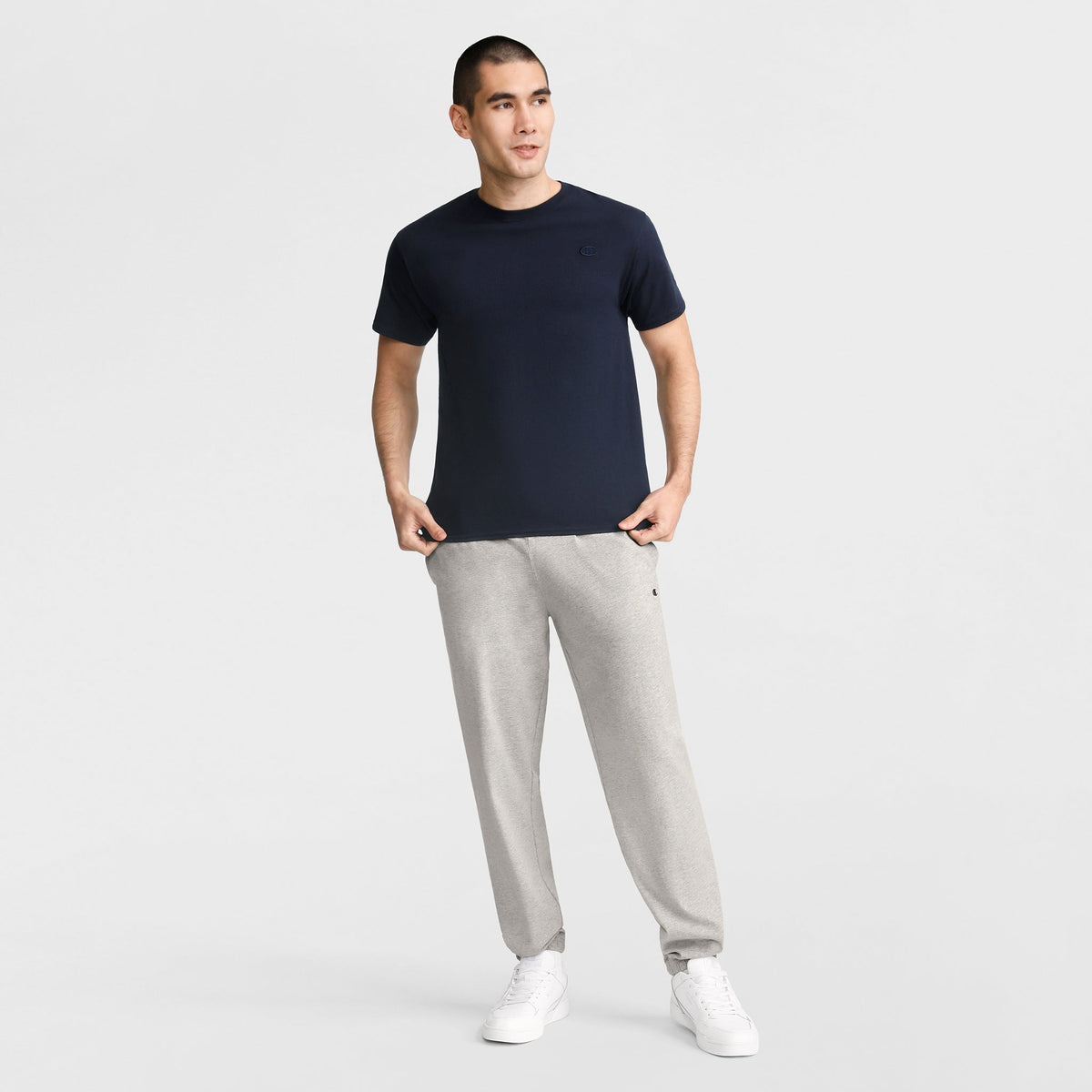 A man stands against a white background, smiling slightly to the side with his hands in his pockets. He wears Champion Lightweight Lounge Pants with cinched hems and C Logo, paired with a navy t-shirt and white sneakers.