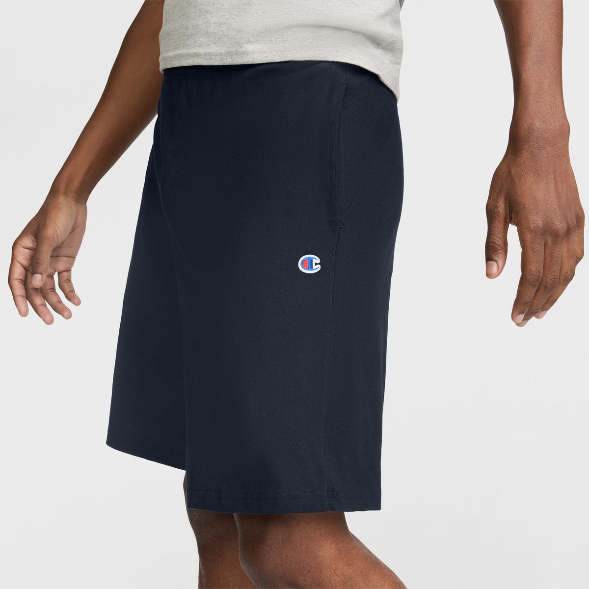 A person is shown from the waist down wearing Champion 9" Lightweight Lounge Shorts with a C Patch Logo in navy blue, crafted from soft cotton jersey, paired with a light gray T-shirt against a plain white background.