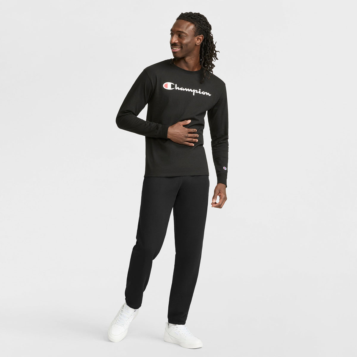 A man smiles while wearing Champion’s Classic Graphic Long-Sleeve T-Shirt with a script logo, paired with black pants and white sneakers. He stands against a plain white background, one hand resting on his stomach as he looks to the side.