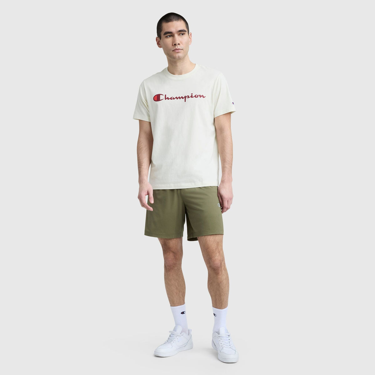 A man faces forward in a white Champion Classic Graphic T-Shirt with Script Logo, olive green shorts, white crew socks, and white sneakers—creating a relaxed streetwear vibe against a simple light gray background.