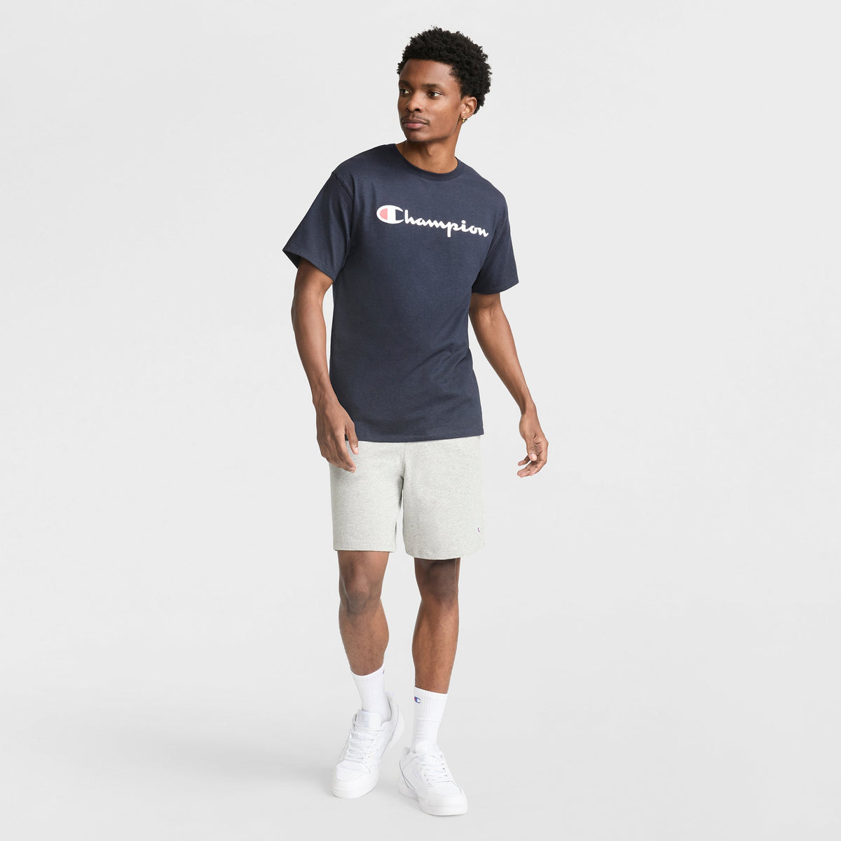A man stands against a white backdrop in a navy Champion Classic Graphic T-Shirt with Script Logo, paired with light gray shorts, white crew socks, and sneakers—capturing an easy streetwear look as he glances sideways with one arm slightly raised.