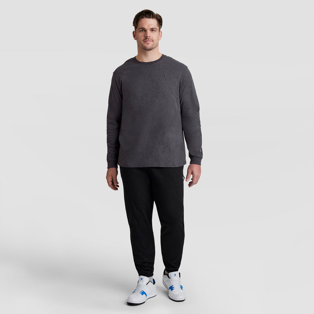 A man wearing a charcoal Classic Long Sleeve T-Shirt, Embroidered C Logo - Big & Tall, black sweatpants, and white sneakers.