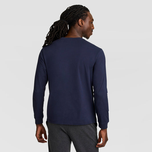 A man with long dreadlocks, seen from behind against a plain light gray background, wears a navy blue Champion Classic Long Sleeve T-Shirt with an embroidered C logo and dark gray pants.
