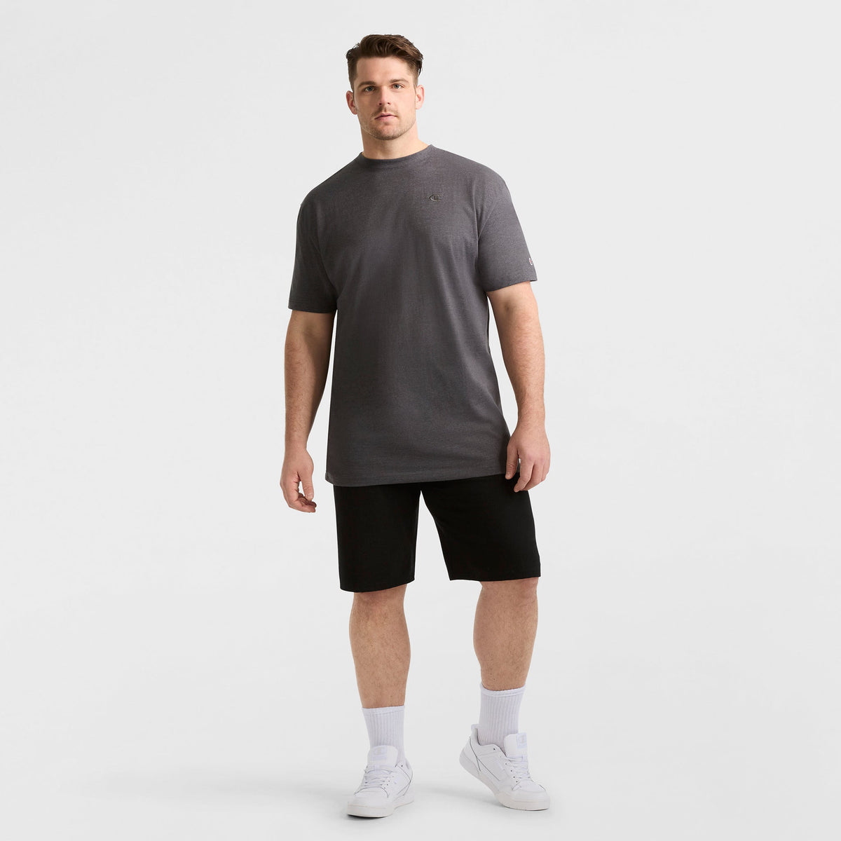 A man faces forward against a white background wearing the Champion Classic T-Shirt, C Logo - Big & Tall in gray, paired with black shorts, white crew socks, and white sneakers. He stands in a relaxed posture.