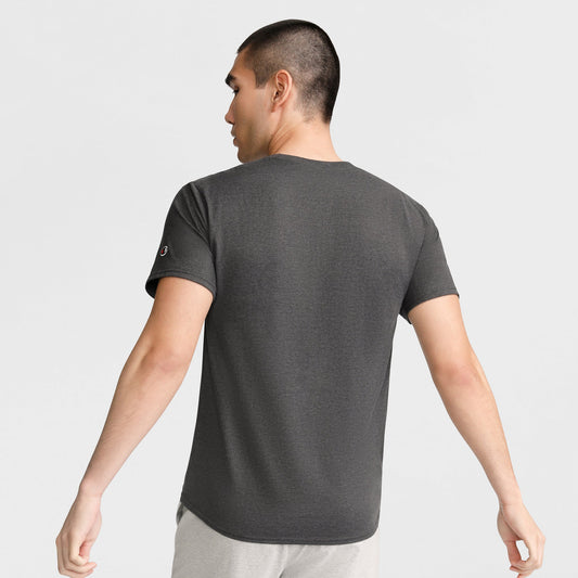 A man with short dark hair stands with his back to the camera, wearing a Champion Classic T-Shirt, C Logo in dark gray and light pants against a plain light background.