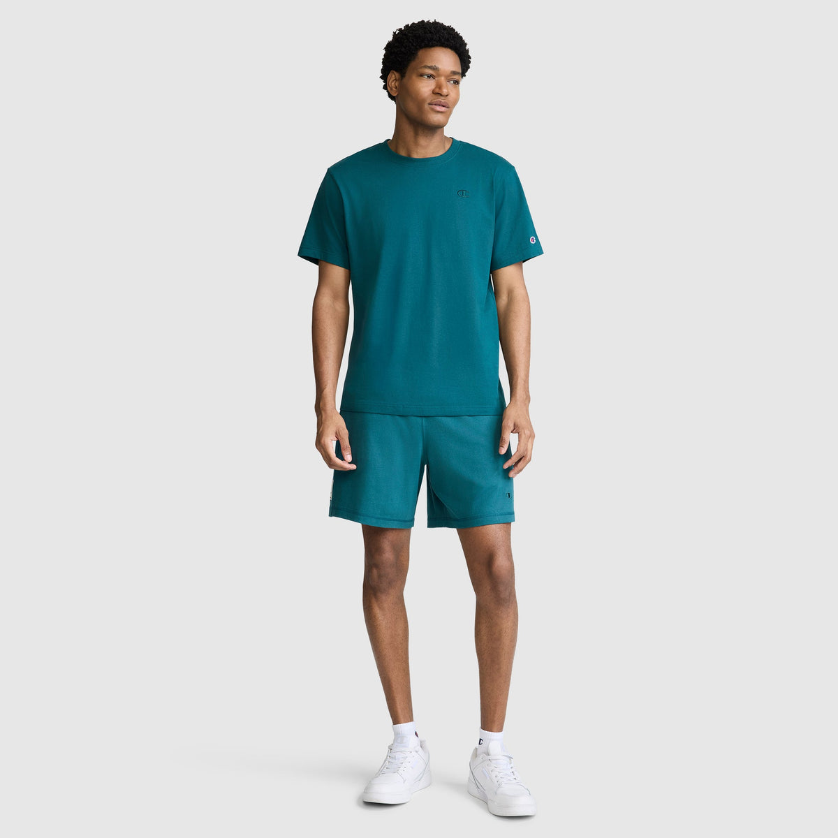 A man stands facing forward wearing a Champion Classic T-Shirt with the C Logo, paired with matching teal shorts and white sneakers against a plain light gray background.