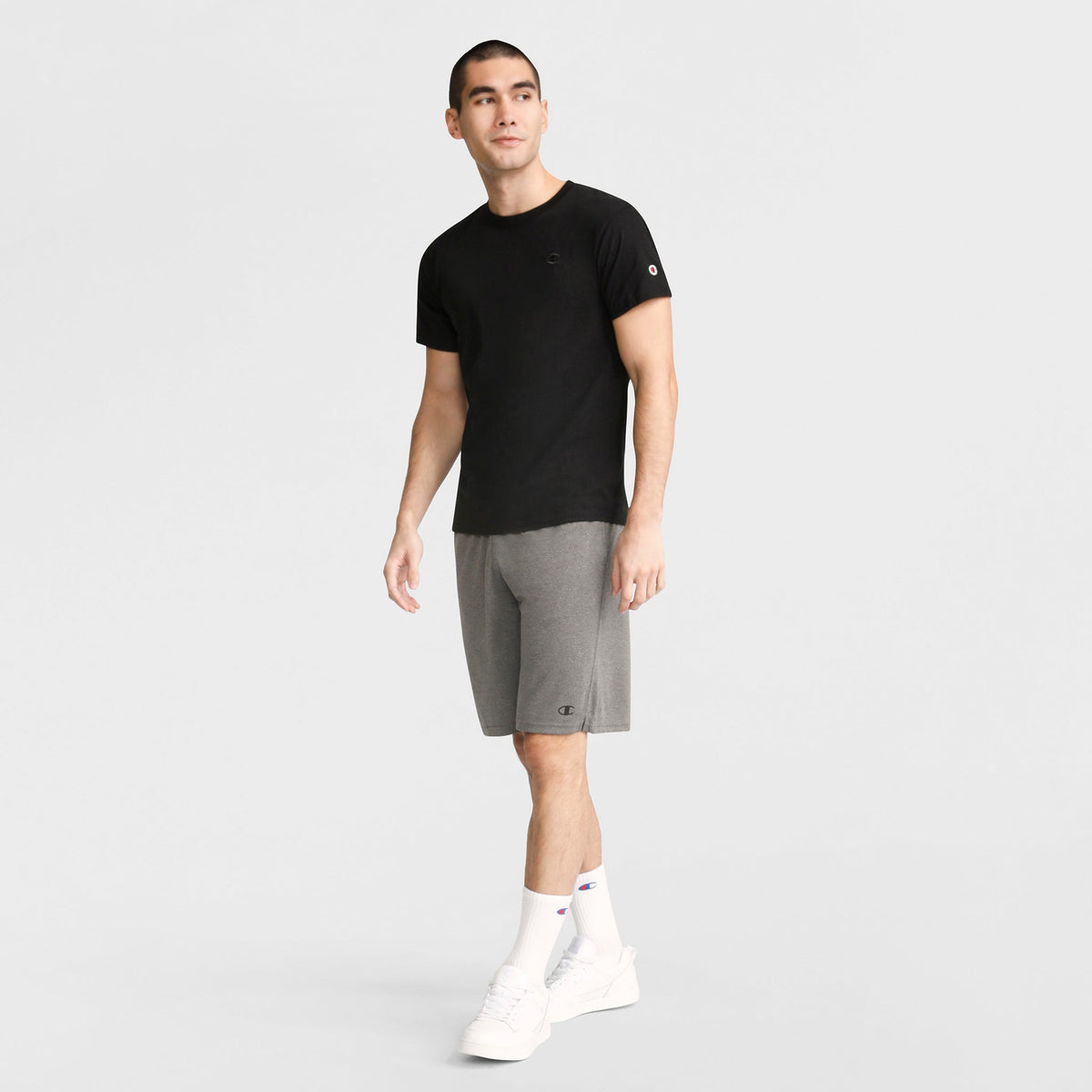 A man stands facing forward in a Champion Classic T-Shirt with C Logo in black, gray shorts, white crew socks, and white sneakers against a plain light background.