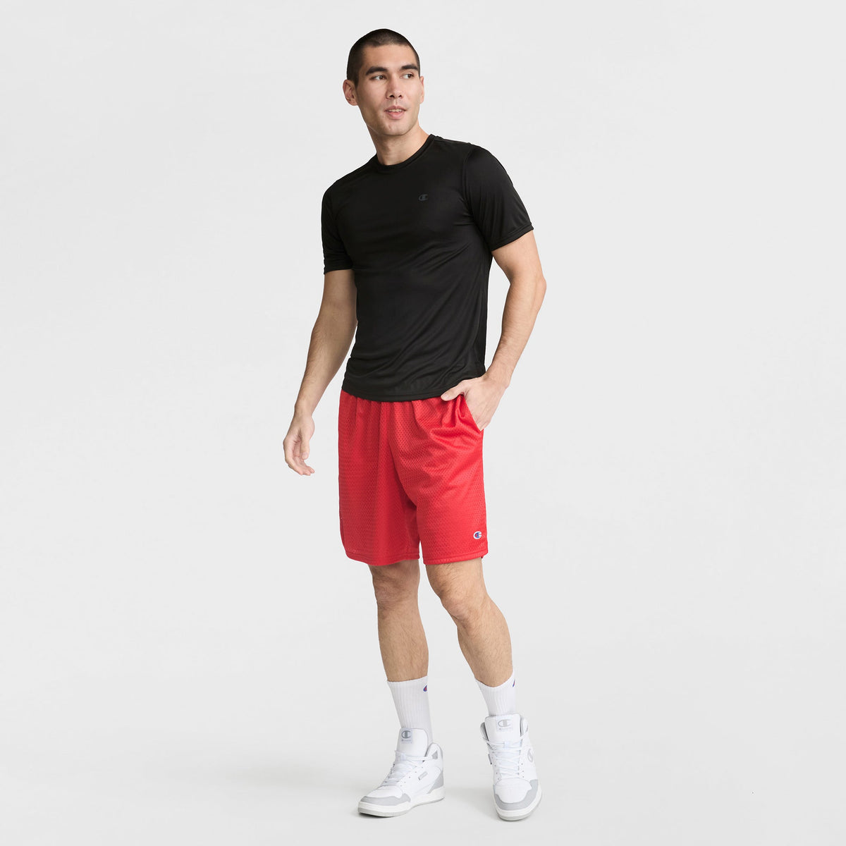 A man stands against a white background wearing a Champion Sport T-Shirt with Reflective C Logo, red athletic shorts, white crew socks, and white sneakers. He has one hand in his pocket and looks slightly to the side.