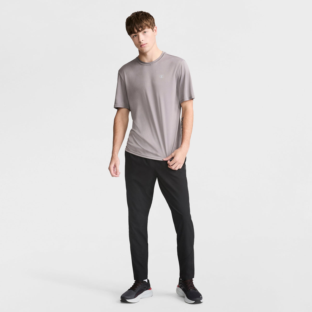 A young man with straight brown hair and a neutral expression stands against a white background, wearing a loose-fitting gray Champion Sport T-Shirt with a reflective C logo, black athletic pants, and black sneakers with white soles.