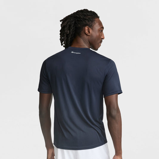 A man with braided hair is seen from the back wearing a navy Champion Sport T-Shirt with a Reflective C Logo and white shorts, set against a plain white background.