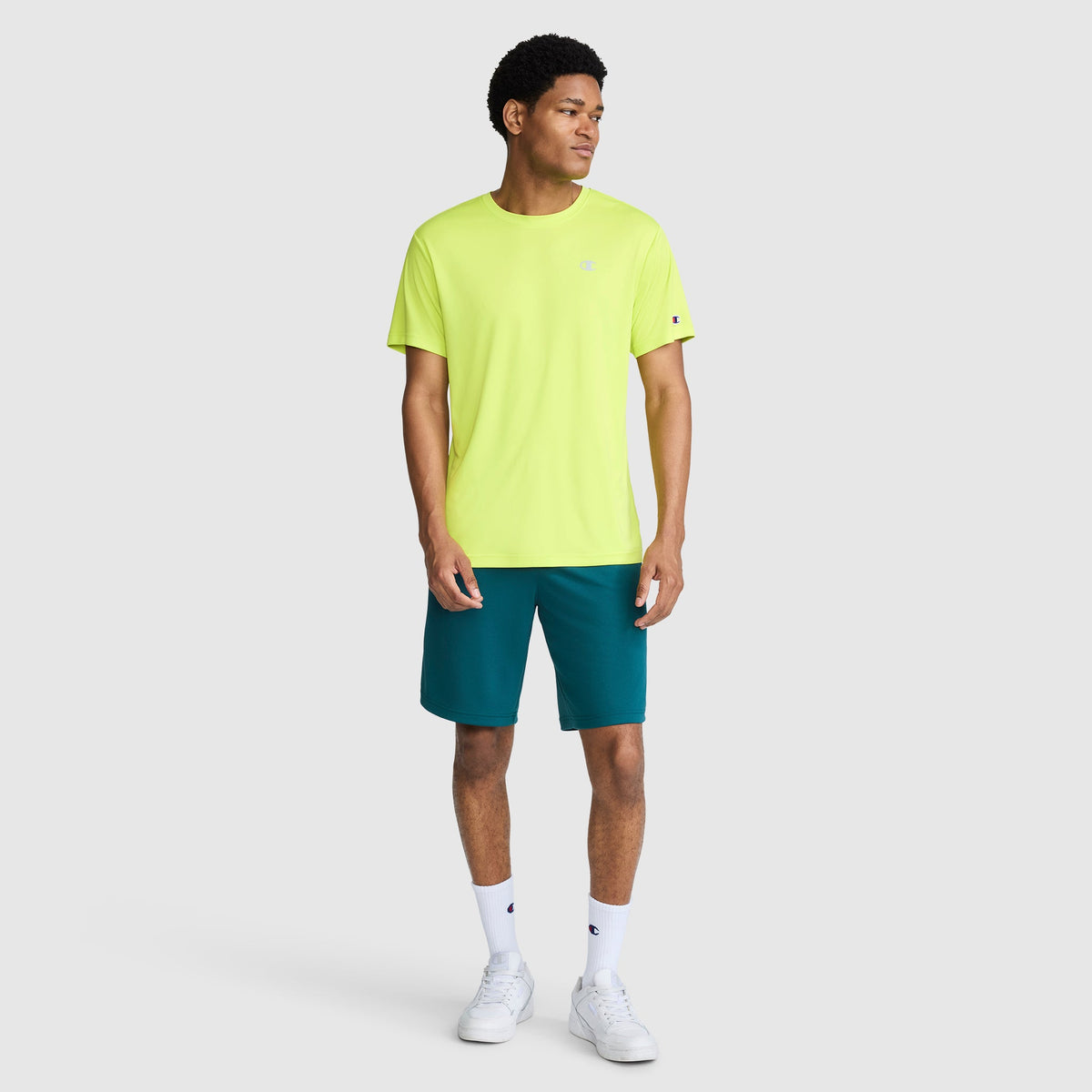 A man stands against a plain light gray background, wearing a bright yellow Champion Sport T-Shirt with a Reflective C Logo, teal shorts, white socks, and white sneakers. He is looking to his left with his arms relaxed by his sides.
