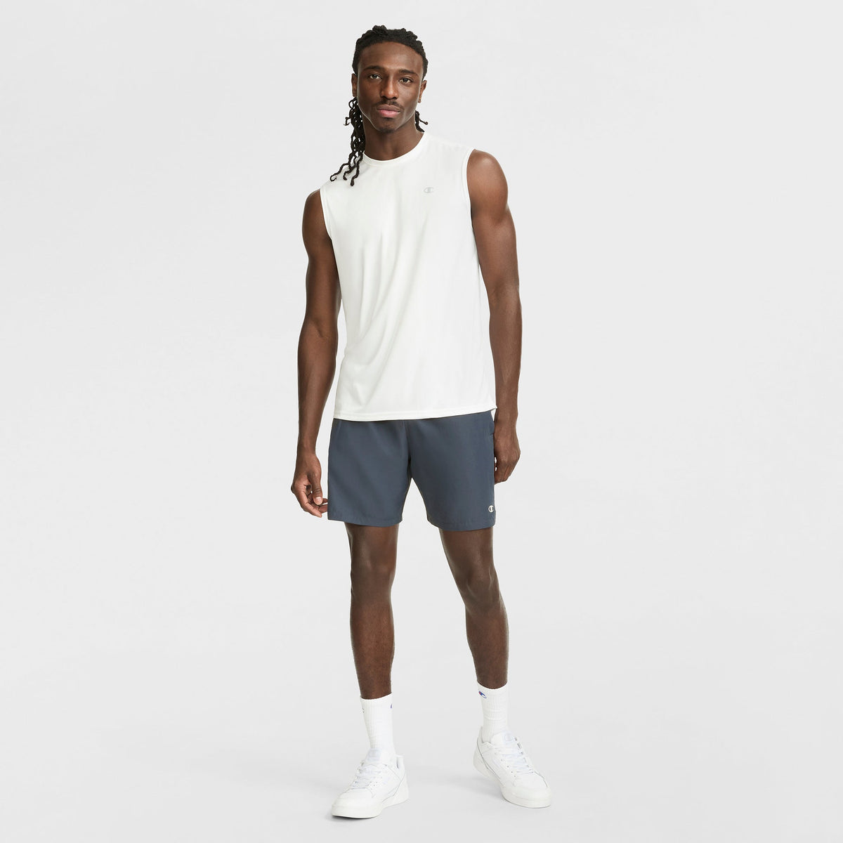 A man with long braided hair stands against a white background wearing a Champion Sport Muscle Tank with Reflective C Logo, dark gray athletic shorts, white crew socks, and white sneakers.