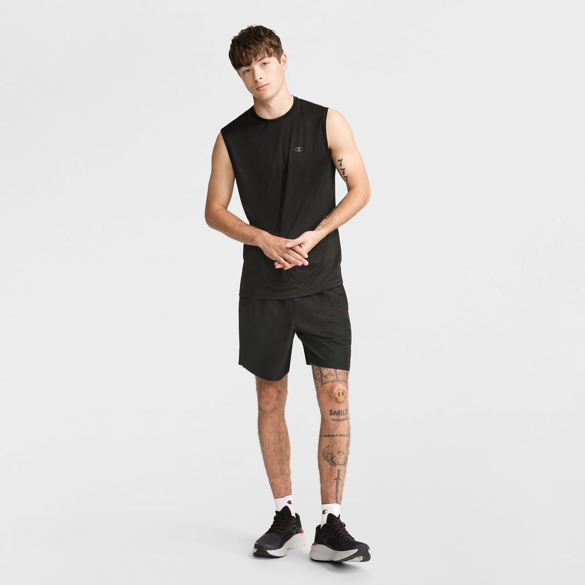 A young man wears the Champion Sport Muscle Tank with Reflective C Logo, paired with black shorts and sneakers. Standing against a white background, he shows off leg tattoos and short hair—an ideal workout shirt for comfort and style.