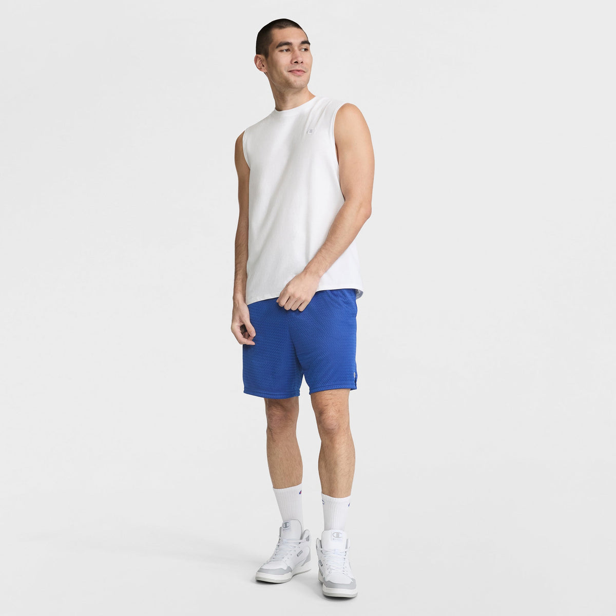 A man smiles and stands against a white background wearing the Champion Muscle Tank, C Logo, paired with blue athletic shorts, white socks, and white sneakers. One hand is by his side while the other touches his shirt.