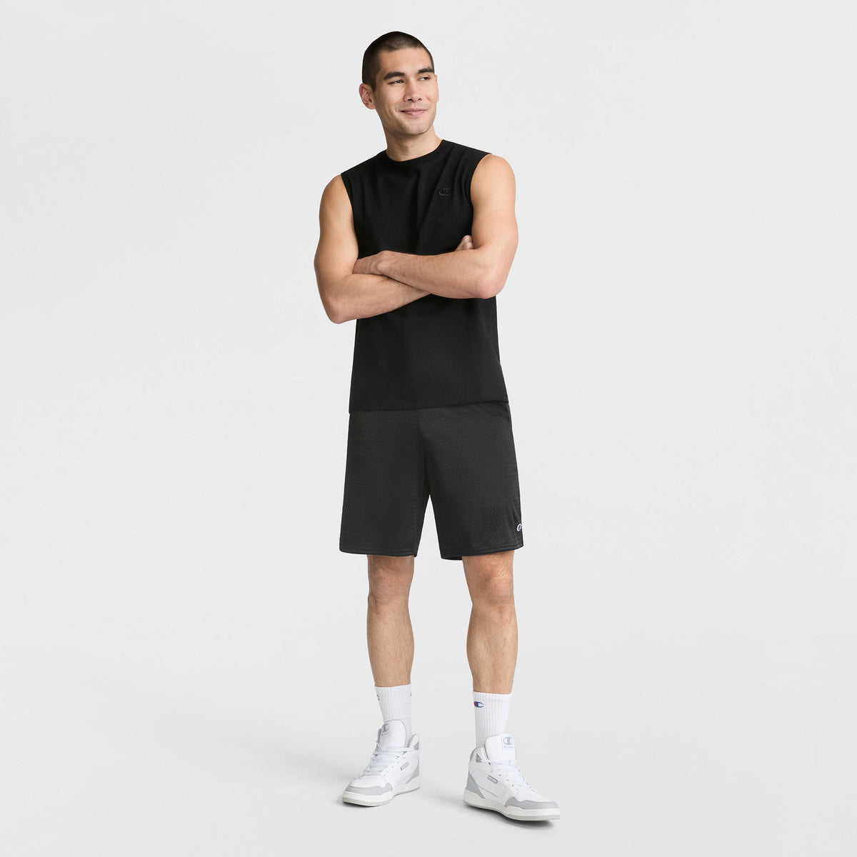 A man stands with arms crossed, wearing a black Champion Muscle Tank, black shorts, white socks, and white sneakers against a plain white background.