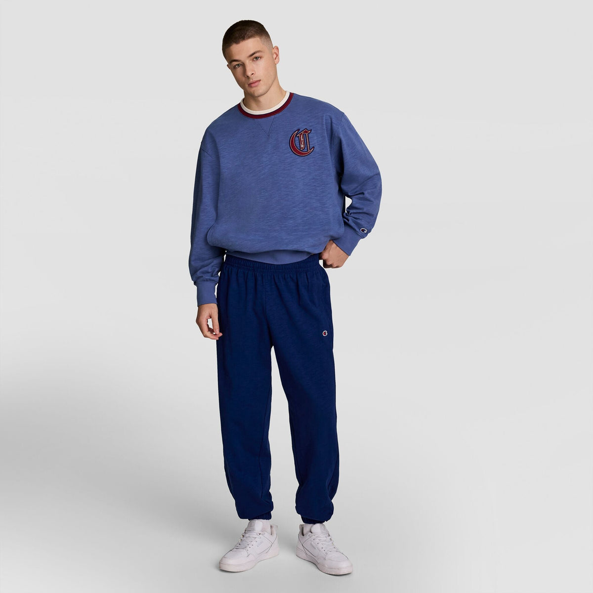 A young man stands against a light grey background wearing a blue Champion crewneck sweatshirt, Modern Retro Washed Pant joggers with C Logo by Champion, and white sneakers.