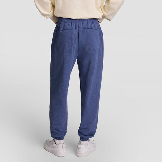 A person is shown from the back wearing a cream sweatshirt, Champion’s Modern Retro Washed Pant, C Logo in blue, and white sneakers, standing on a plain light background.