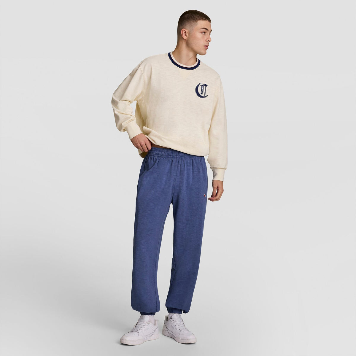 A young man stands against a light gray background wearing Champion’s cream-colored sweatshirt, Modern Retro Washed Pant with C Logo, and white sneakers. He looks to the side with one hand in his pocket.
