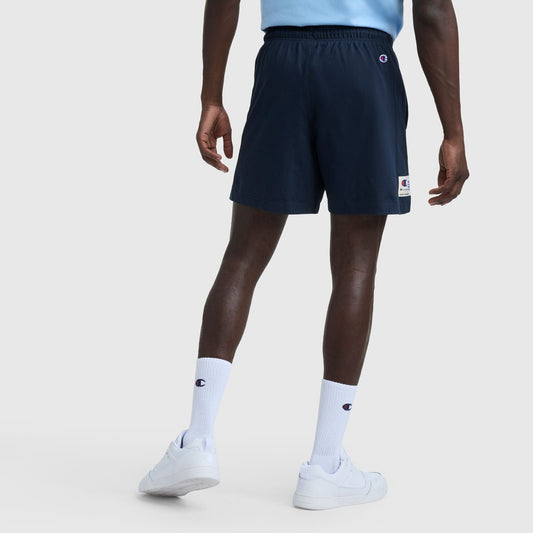 A person faces away, wearing a light blue heritage cotton jersey shirt, Champion 7″ Jersey Shorts, Script Logo in navy, white crew socks with Champion script, and white sneakers against a plain light background.