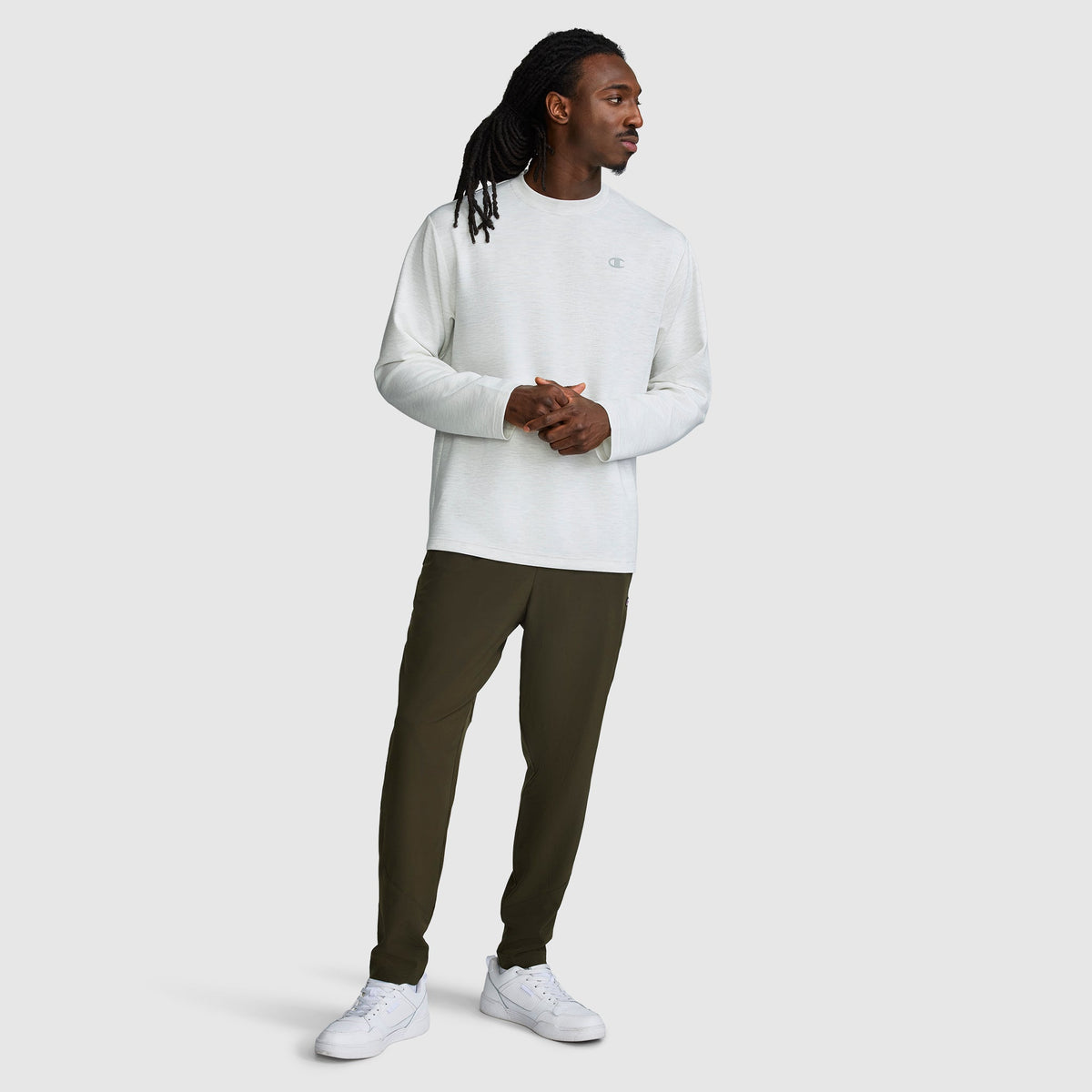 A man stands against a plain background wearing a Champion Sport Long Sleeve Crewneck, C Logo in white, paired with dark green pants and white sneakers. He looks to the side with his hands loosely clasped.